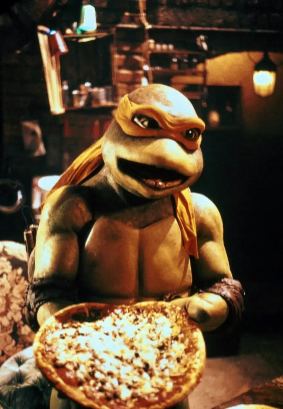 A Teenage Mutant Ninja Turtle with an orange mask holds a pizza, smiling in a dimly lit, cozy room with brick walls and hanging lights.