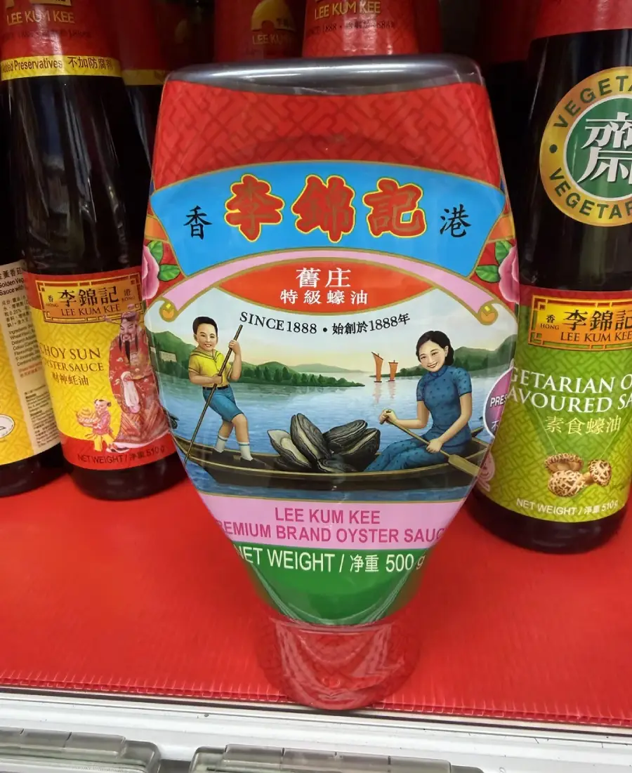 A bottle of Lee Kum Kee Premium Brand Oyster Sauce is on a shelf, featuring an illustration of two people collecting oysters by a river, surrounded by other bottles.