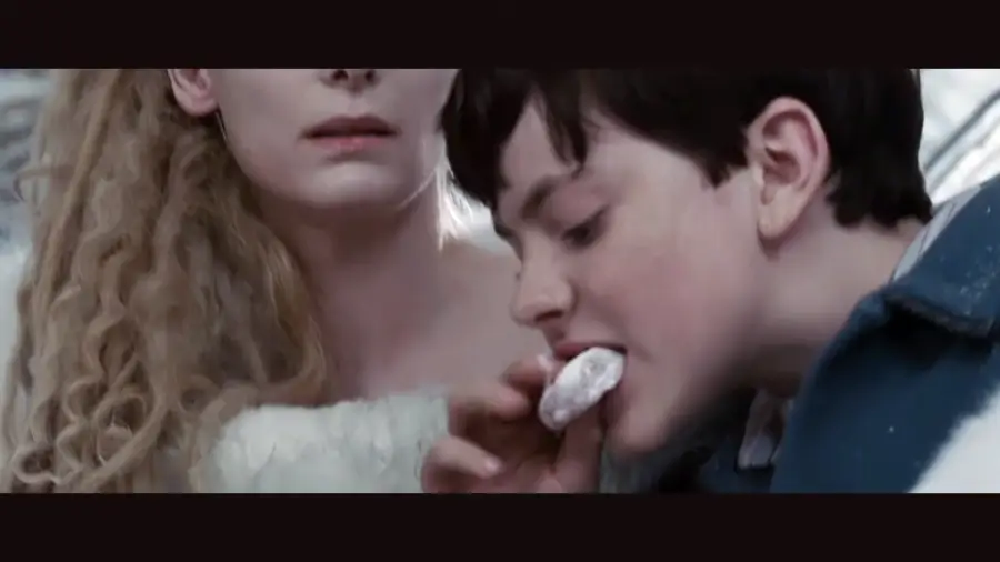 A woman with long, curly blonde hair looks on as a boy with short dark hair takes a bite of a powdered treat she is holding. Both are dressed in light-colored clothing.