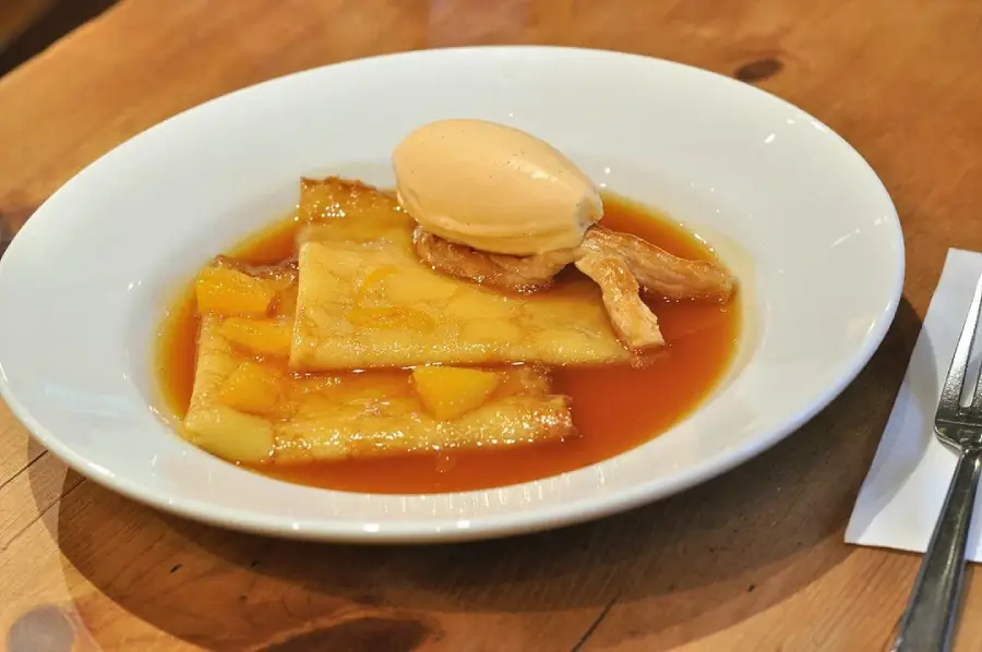 A white plate with crepes in orange sauce, topped with a scoop of ice cream, sits on a wooden table next to a fork and napkin.