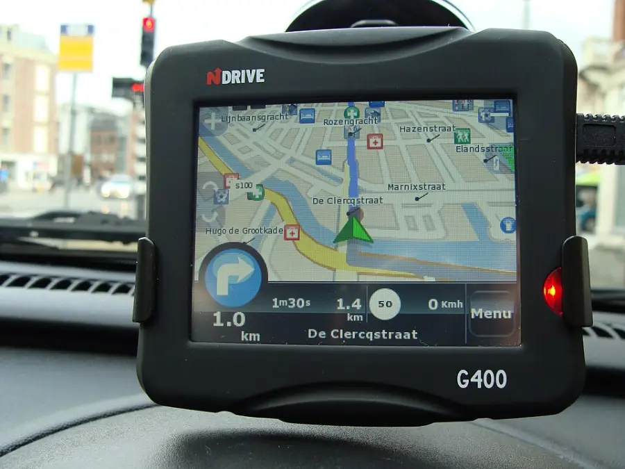 A GPS device mounted in a car displays a map with directions, showing street names and a route marked in blue. The screen indicates a left turn ahead and distance to the next maneuver.