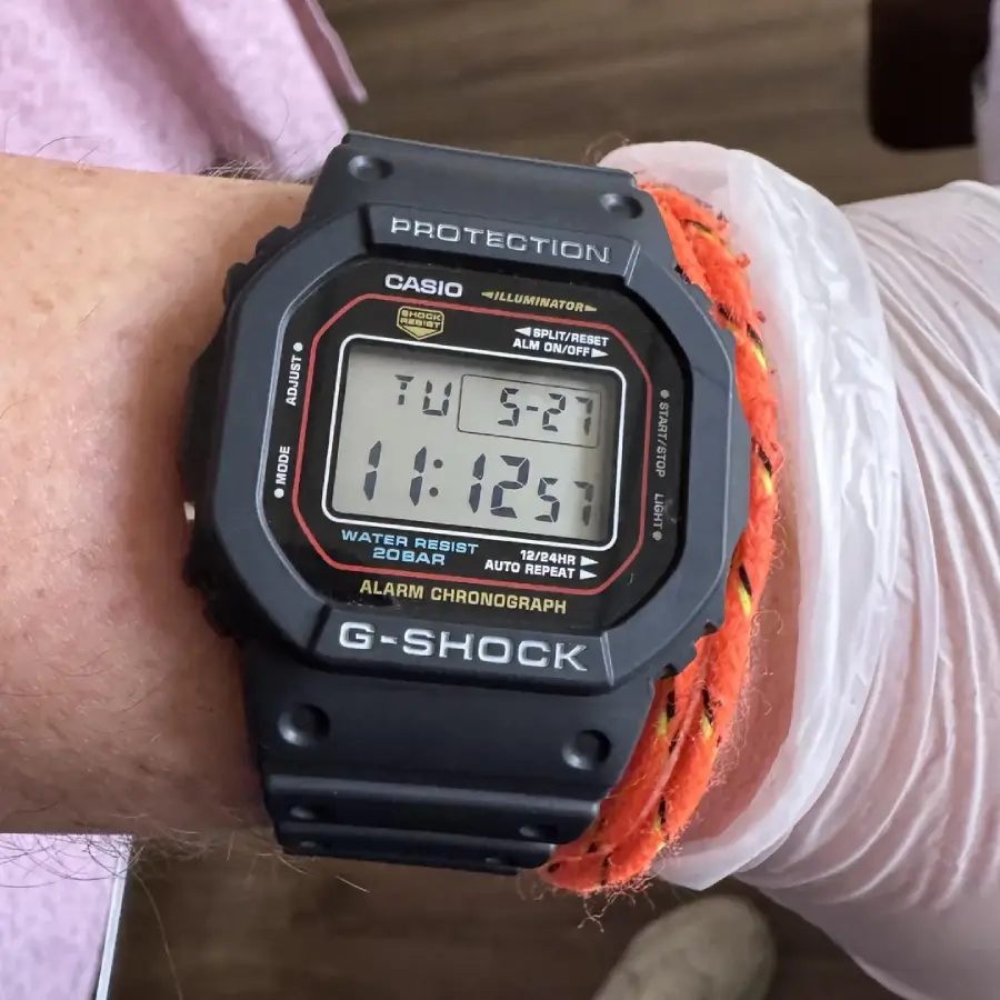 A wrist with a black Casio G-Shock digital watch displaying the time 11:12:51 and the date as Tuesday, 5/21. The person also wears an orange braided bracelet and a light glove.