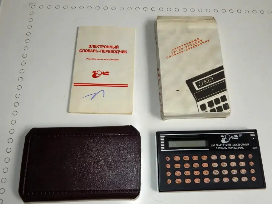 A Russian electronic dictionary-translator set with a manual, original box, brown case, and device featuring small orange and white buttons on a black background, laid out on a white surface.