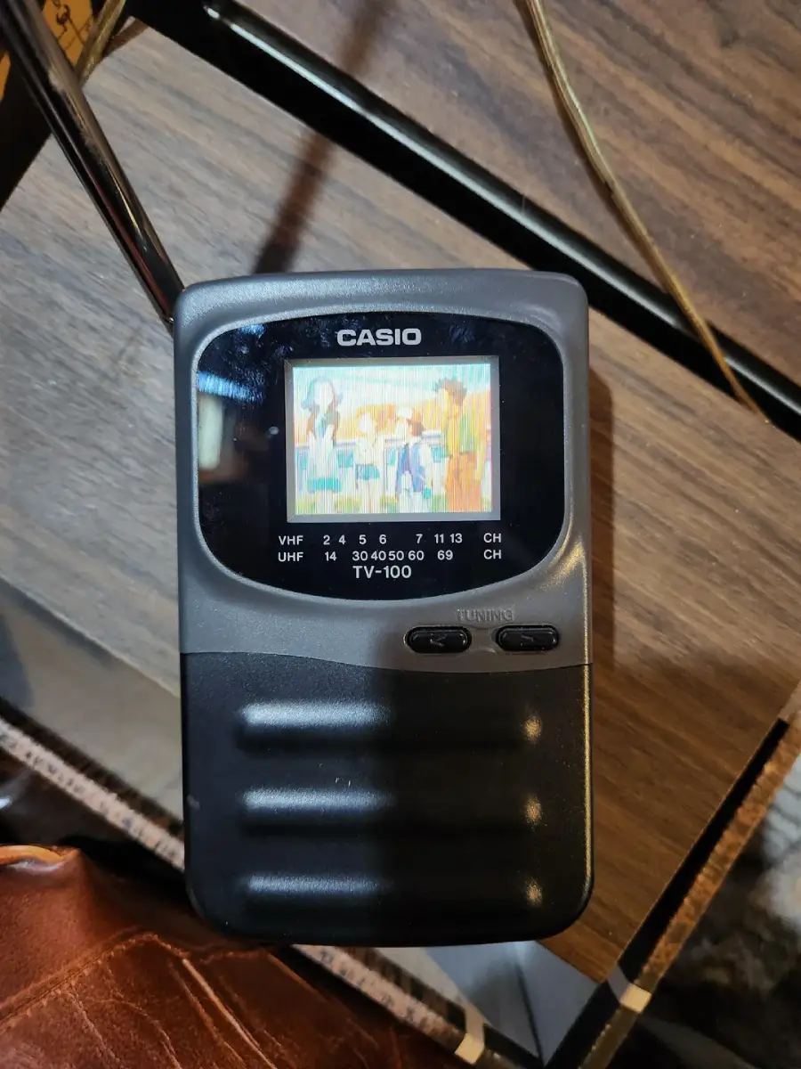 A black Casio TV-100 portable television sits on a wooden table, displaying a colorful cartoon on its small screen. Tuning dials and controls are visible below the screen.