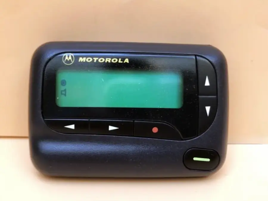 A black Motorola pager with a small screen and several buttons, including directional arrows and a green button, set against a plain beige background.