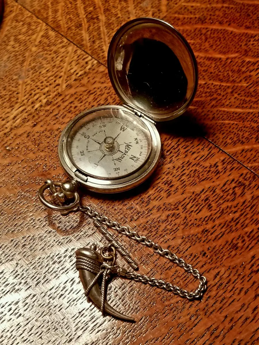A silver pocket compass with its lid open sits on a wooden surface. Attached to its chain is a pendant shaped like a helmet with a feathered plume.