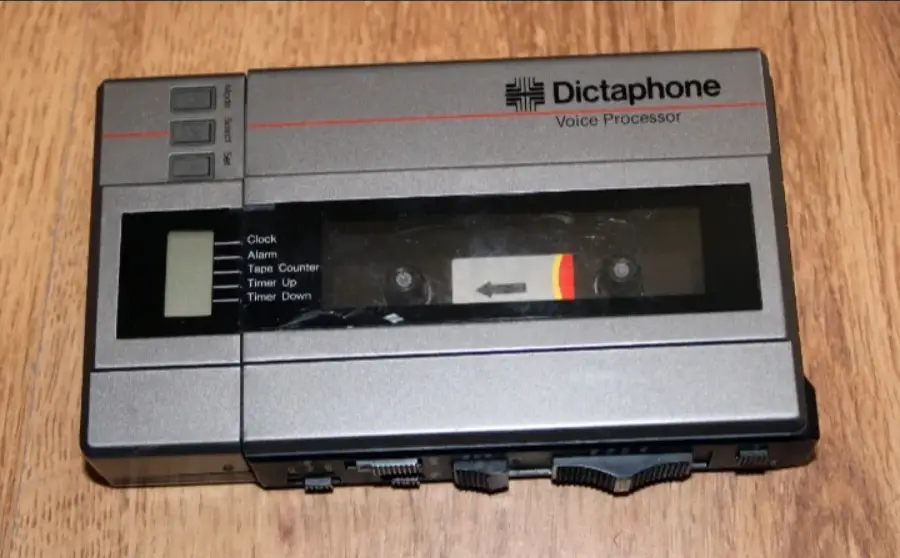 A silver and black Dictaphone voice processor with buttons, a small digital display, and a visible cassette tape inside, placed on a wooden surface.