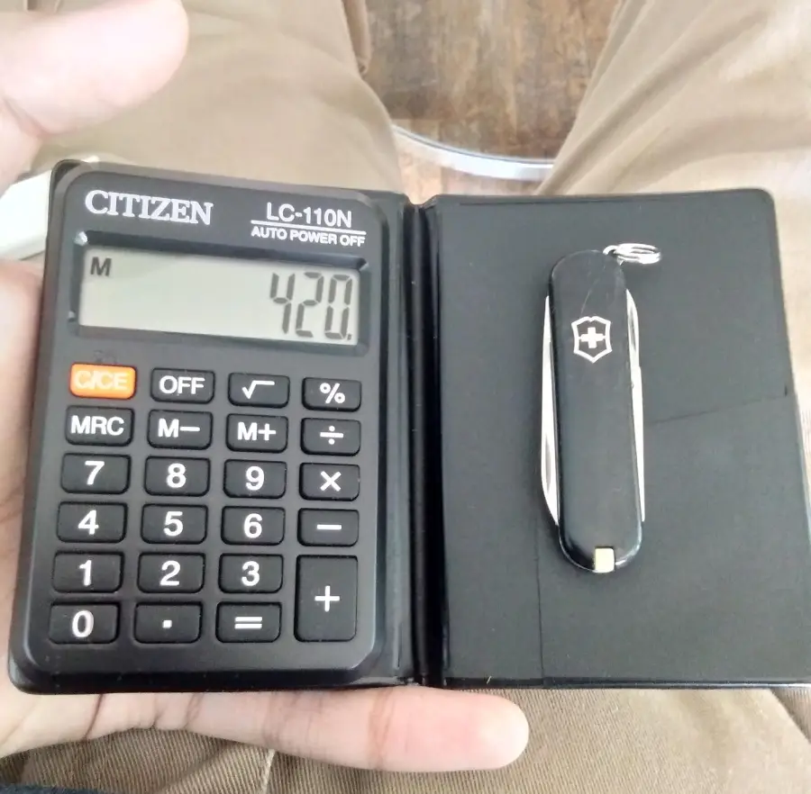 A hand holds an open black case containing a Citizen LC-110N calculator displaying "420" on the screen and a black Swiss Army pocket knife beside it. Brown pants and a wooden floor are visible in the background.