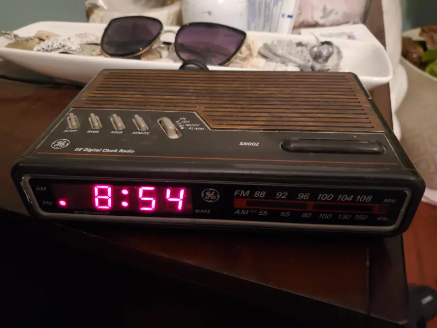 A vintage GE digital clock radio displays the time 8:54 PM in red LED numbers. The radio has buttons and dials on top, with sunglasses and a tray of items in the background.
