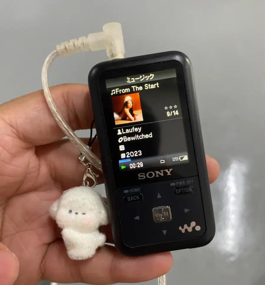 A hand holds a black Sony Walkman music player with a white plush keychain attached. The screen displays the song "From The Start" by Laufey from the album "Bewitched." An earphone is plugged in.