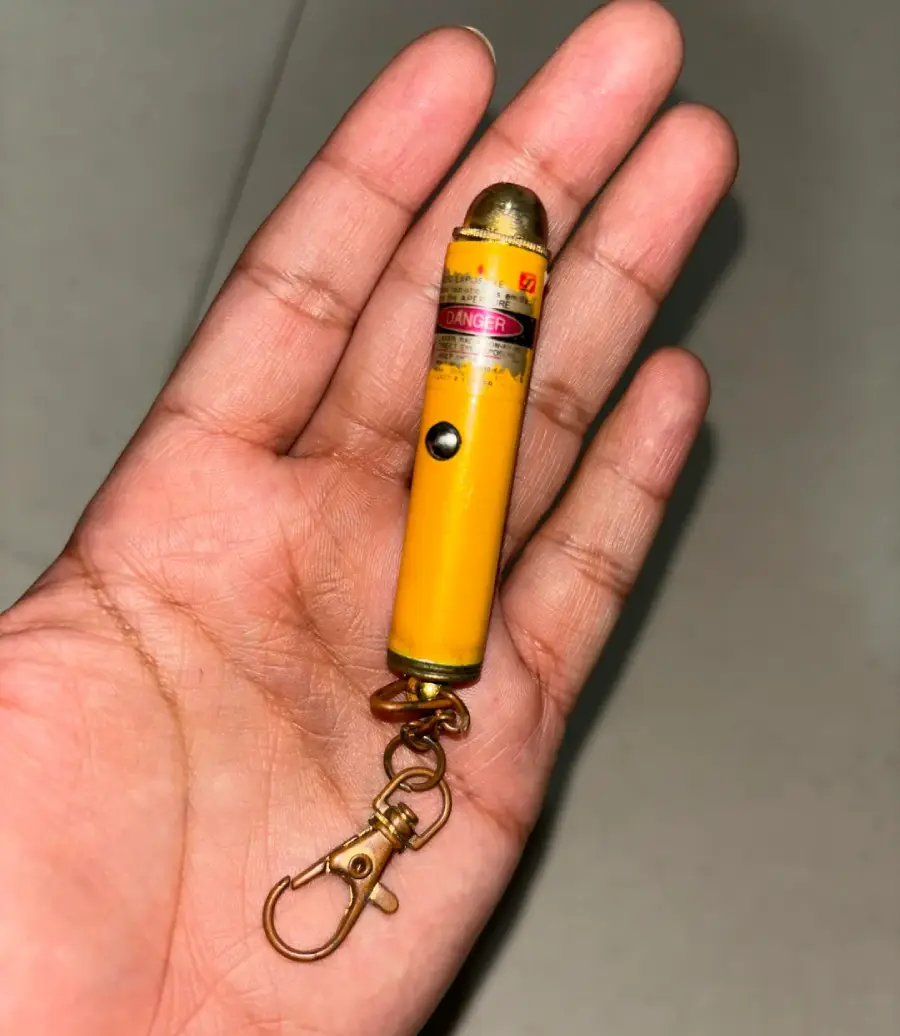 A hand holding a yellow laser pointer keychain with a metal clasp and a warning label near the top.