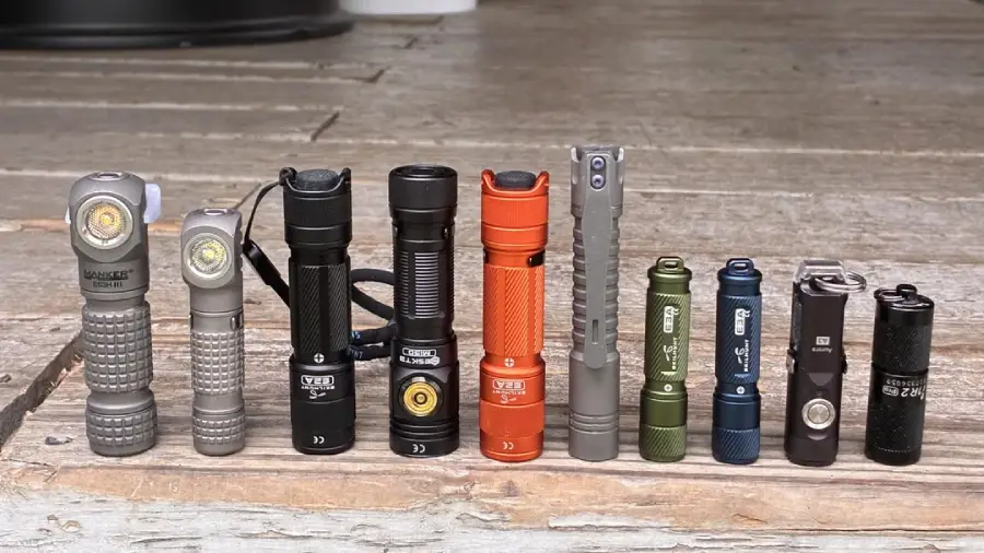 A row of ten different flashlights in various colors and sizes is lined up on a wooden surface, with finishes including silver, black, orange, green, blue, and gray.