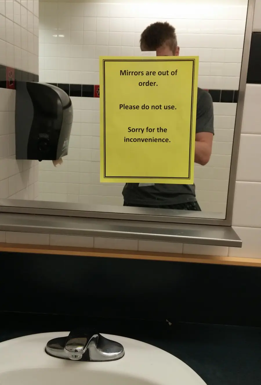 A yellow sign on a mirror reads: "Mirrors are out of order. Please do not use. Sorry for the inconvenience." A person is partially visible in the reflection near a sink and soap dispenser.