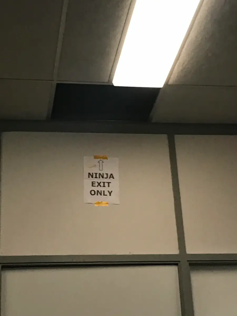 A sign on a wall reads "NINJA EXIT ONLY" with an arrow pointing up to an open ceiling tile above it, near fluorescent lights.