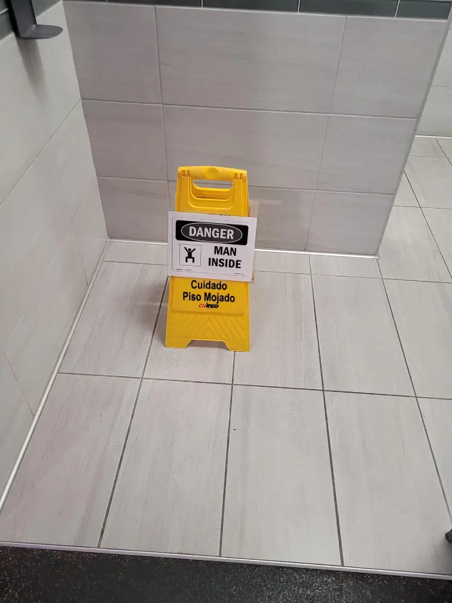 A yellow caution sign on a tiled floor reads "DANGER MAN INSIDE" on top and "Cuidado Piso Mojado" below, indicating a wet floor in both English and Spanish. The sign is placed near a wall in a restroom.