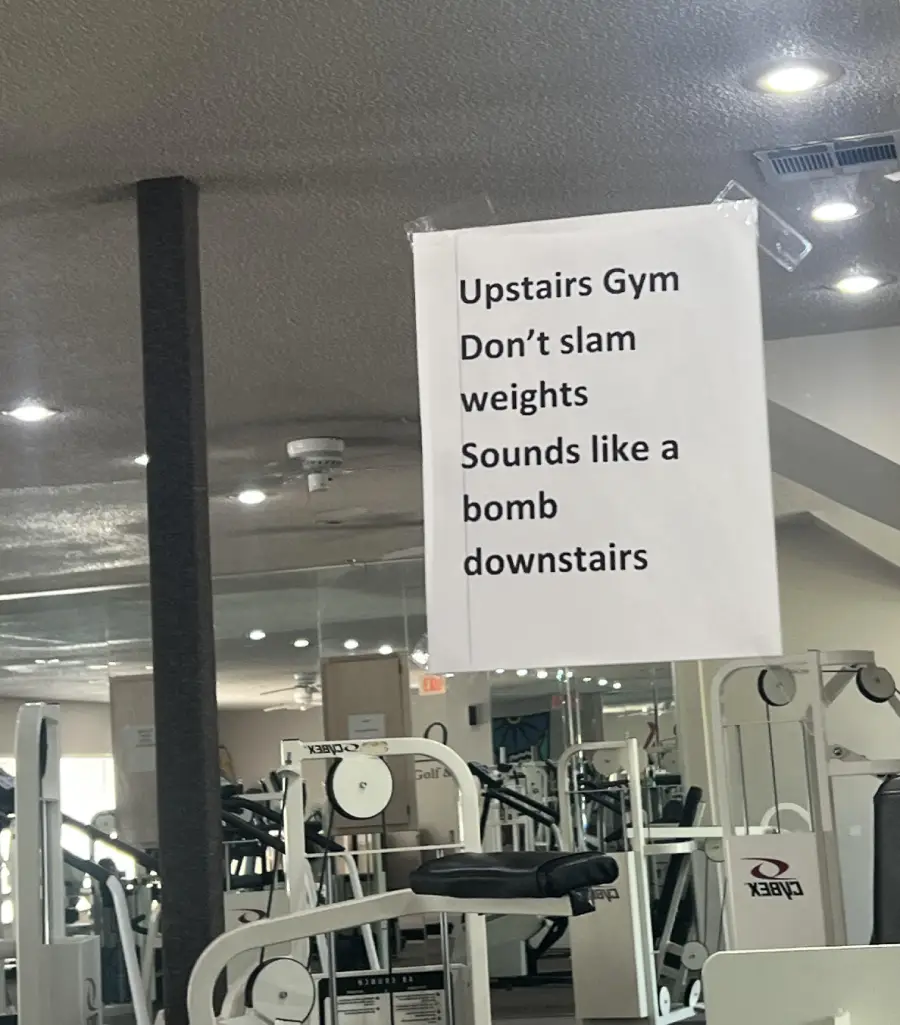 A gym sign reads: "Upstairs Gym. Don't slam weights. Sounds like a bomb downstairs." Gym equipment and mirrors are visible in the background.