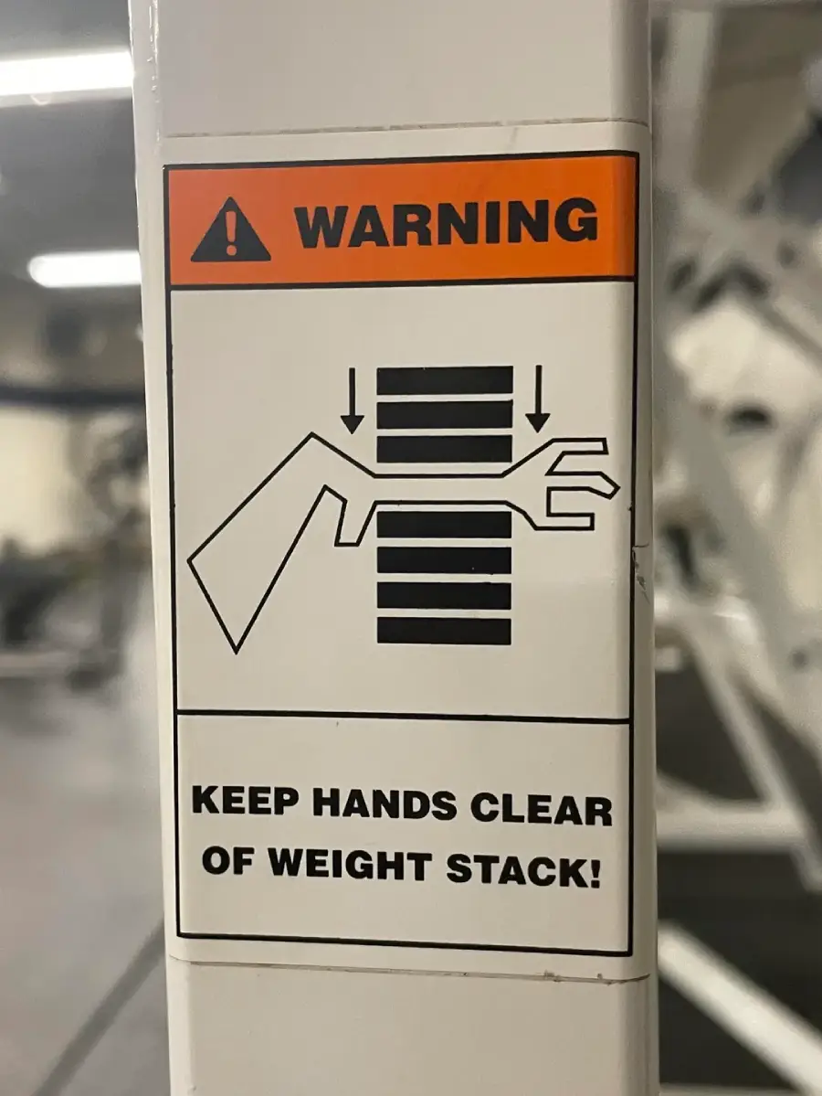 Warning sign on gym equipment shows a hand being pinched by weights, with text: "Keep hands clear of weight stack!" and an exclamation icon above.