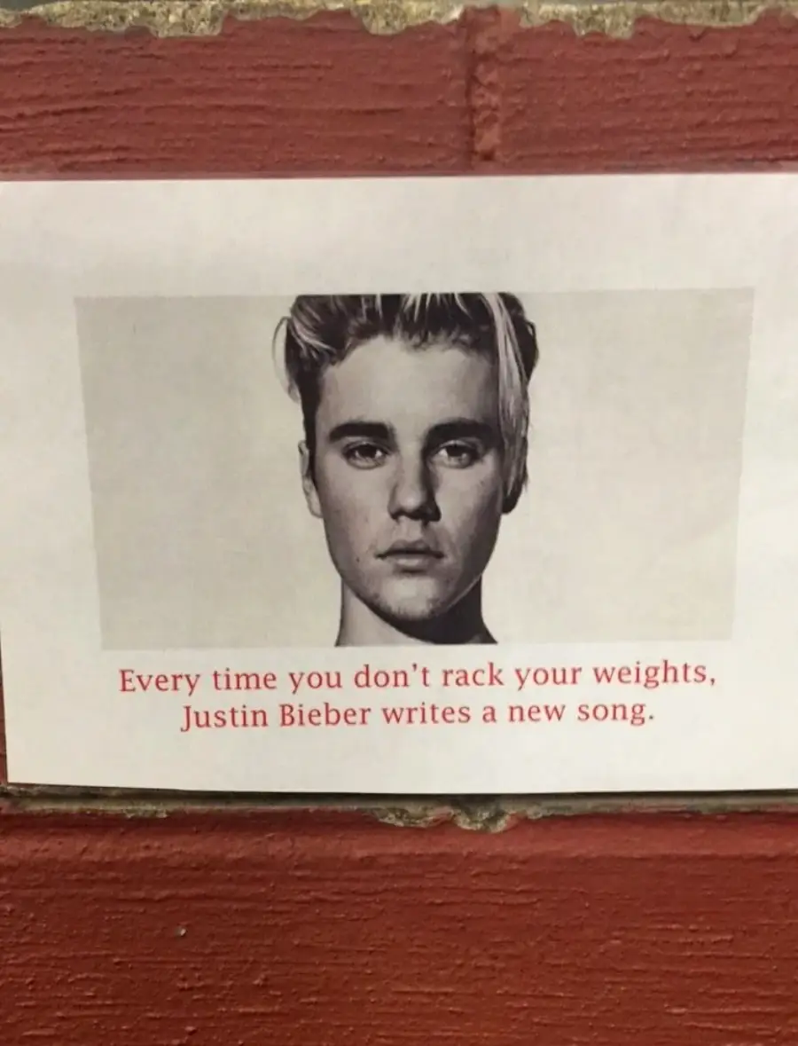 A black-and-white photo of a young man is above text that reads: "Every time you don’t rack your weights, Justin Bieber writes a new song." The sign is posted on a brick wall.