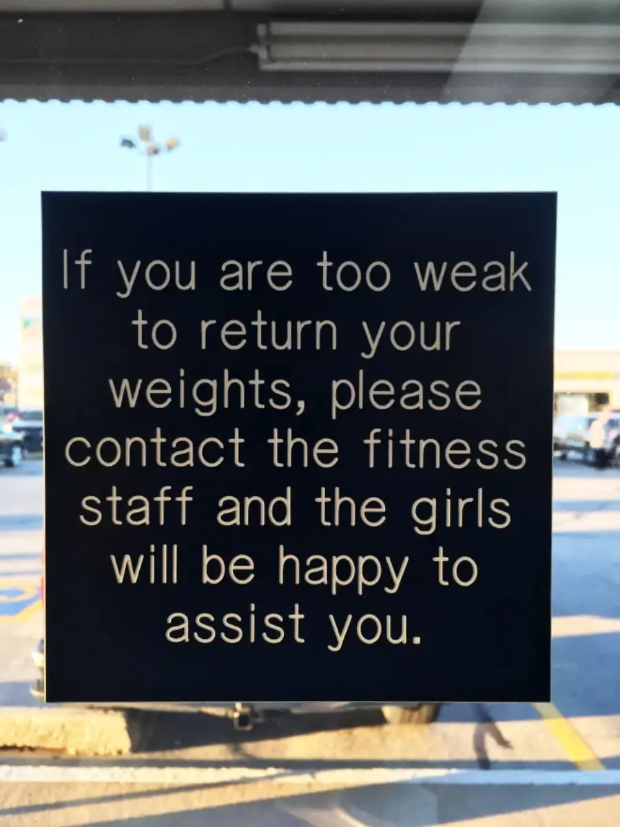 A sign reads: "If you are too weak to return your weights, please contact the fitness staff and the girls will be happy to assist you." Cars are visible outside through the window.