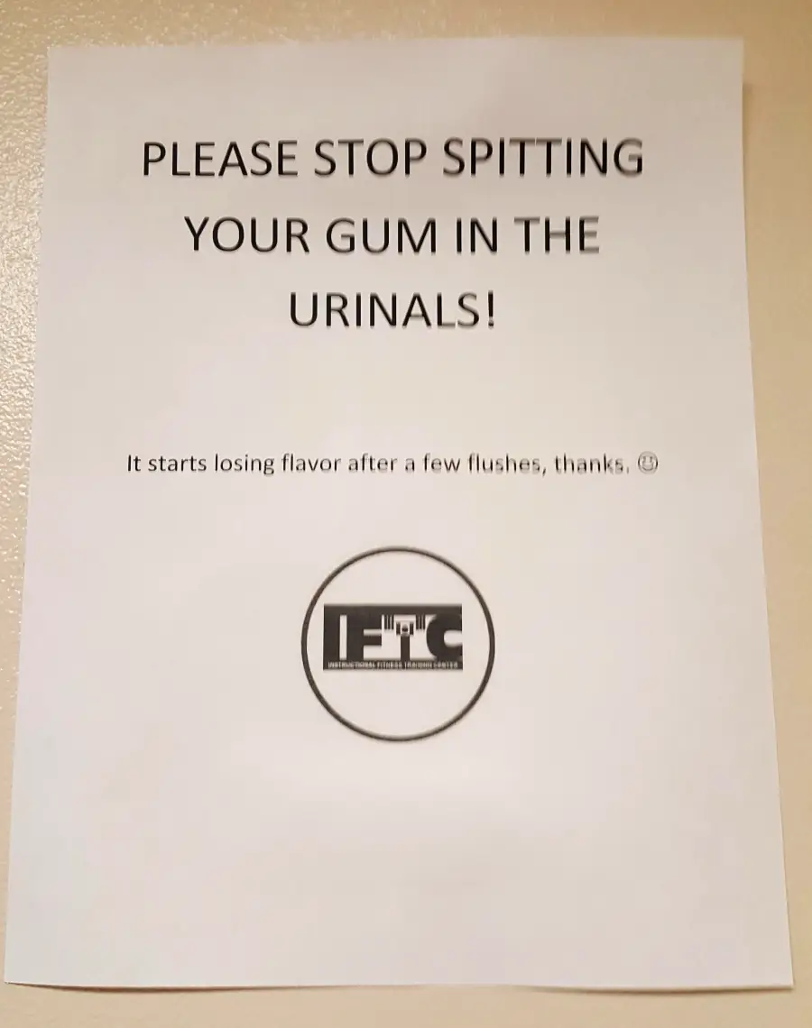 A sign reads: "PLEASE STOP SPITTING YOUR GUM IN THE URINALS! It starts losing flavor after a few flushes, thanks." There is a circular logo at the bottom.