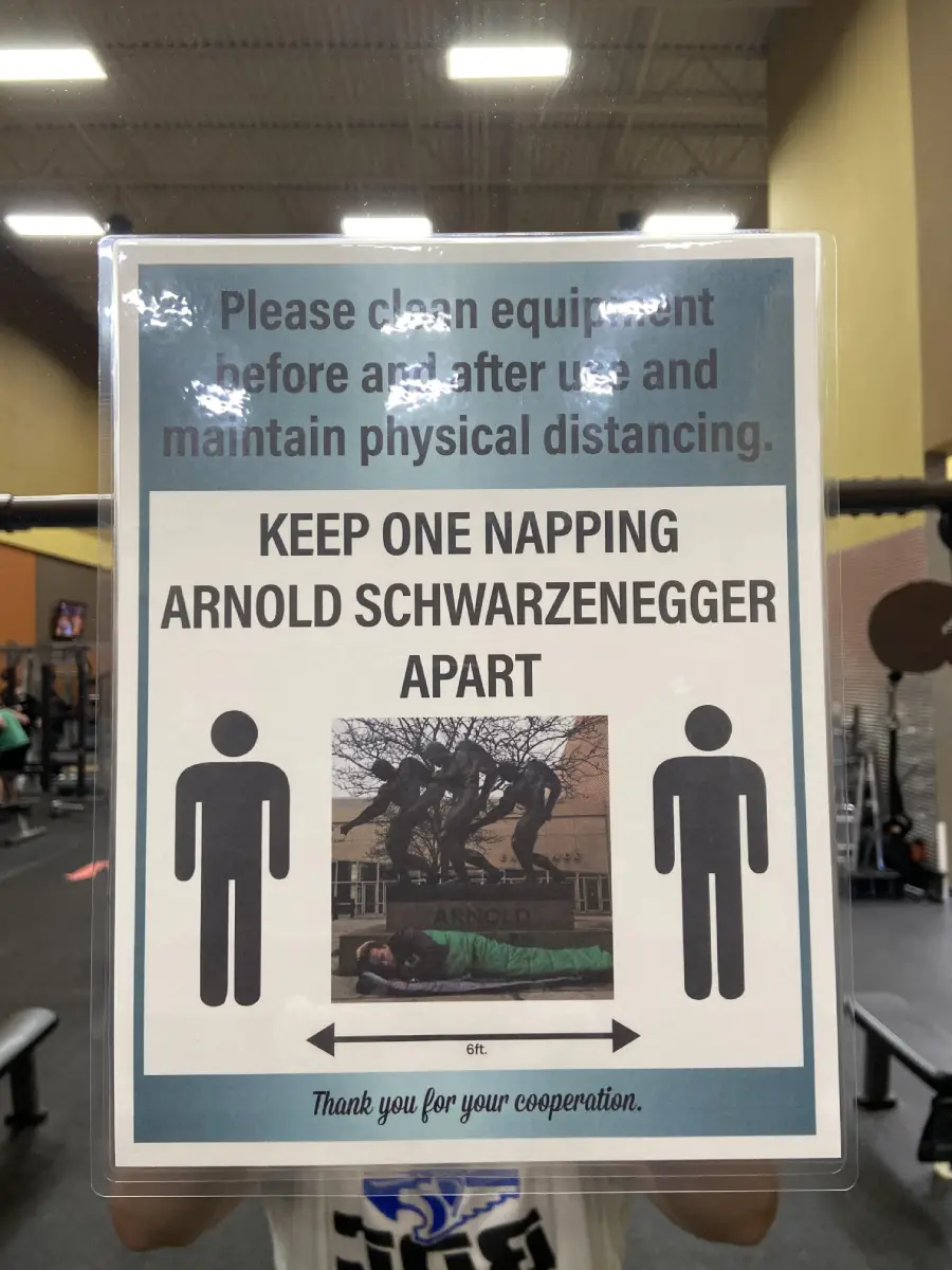 A gym sign reads, "Keep one napping Arnold Schwarzenegger apart," with a photo of Arnold lying on a bench and icons showing a 6 ft distance, reminding people to maintain physical distancing.
