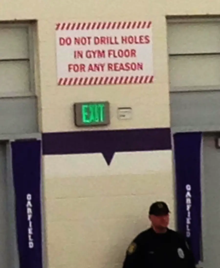 A red and white sign above an exit reads, "DO NOT DRILL HOLES IN GYM FLOOR FOR ANY REASON." A security guard stands below the sign.