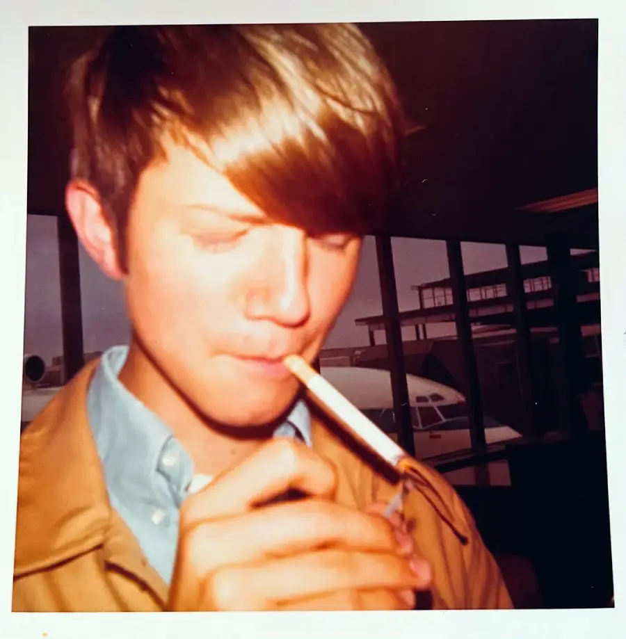 A young person with brown hair lights a cigarette indoors near large windows, with an airplane visible outside in the background.