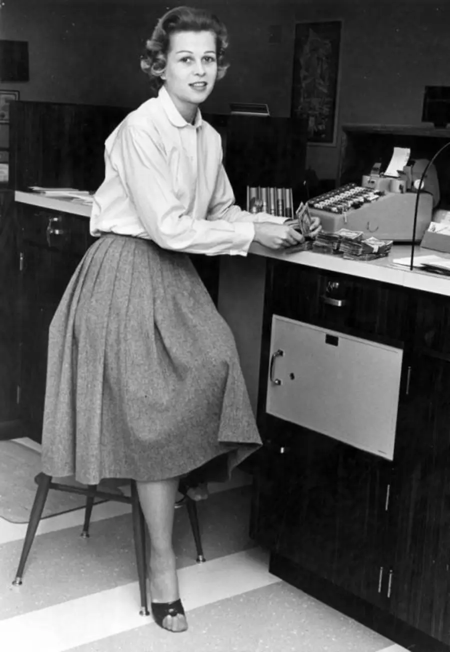 A woman in a blouse and long skirt sits on a stool at a counter, using a vintage cash register. She looks at the camera, and the setting appears to be a mid-20th-century office or bank.