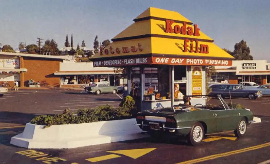 A vintage drive-thru Kodak Film booth offers photo finishing services in a parking lot, with a classic green convertible car parked beside it and shops in the background.