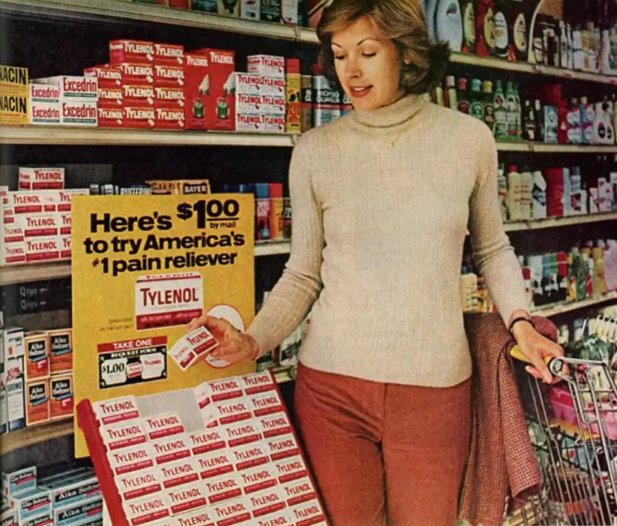 A woman in a grocery store picks up a Tylenol sample from a display offering a $1.00 mail-in trial. The shelves behind her are stocked with various medicines, and she has one hand on her shopping cart.