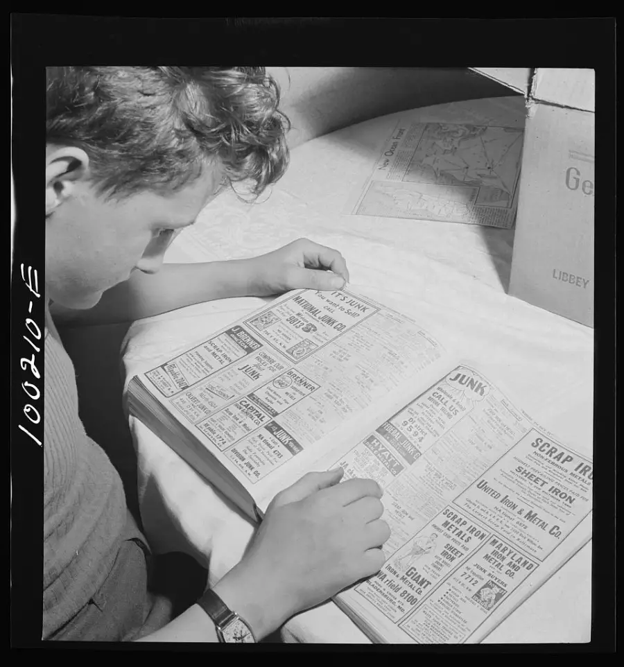 A person sits at a table, looking through a telephone directory or classified ads book. The page features various advertisements for services like scrap iron and junk collection. A cardboard box and paper are nearby.