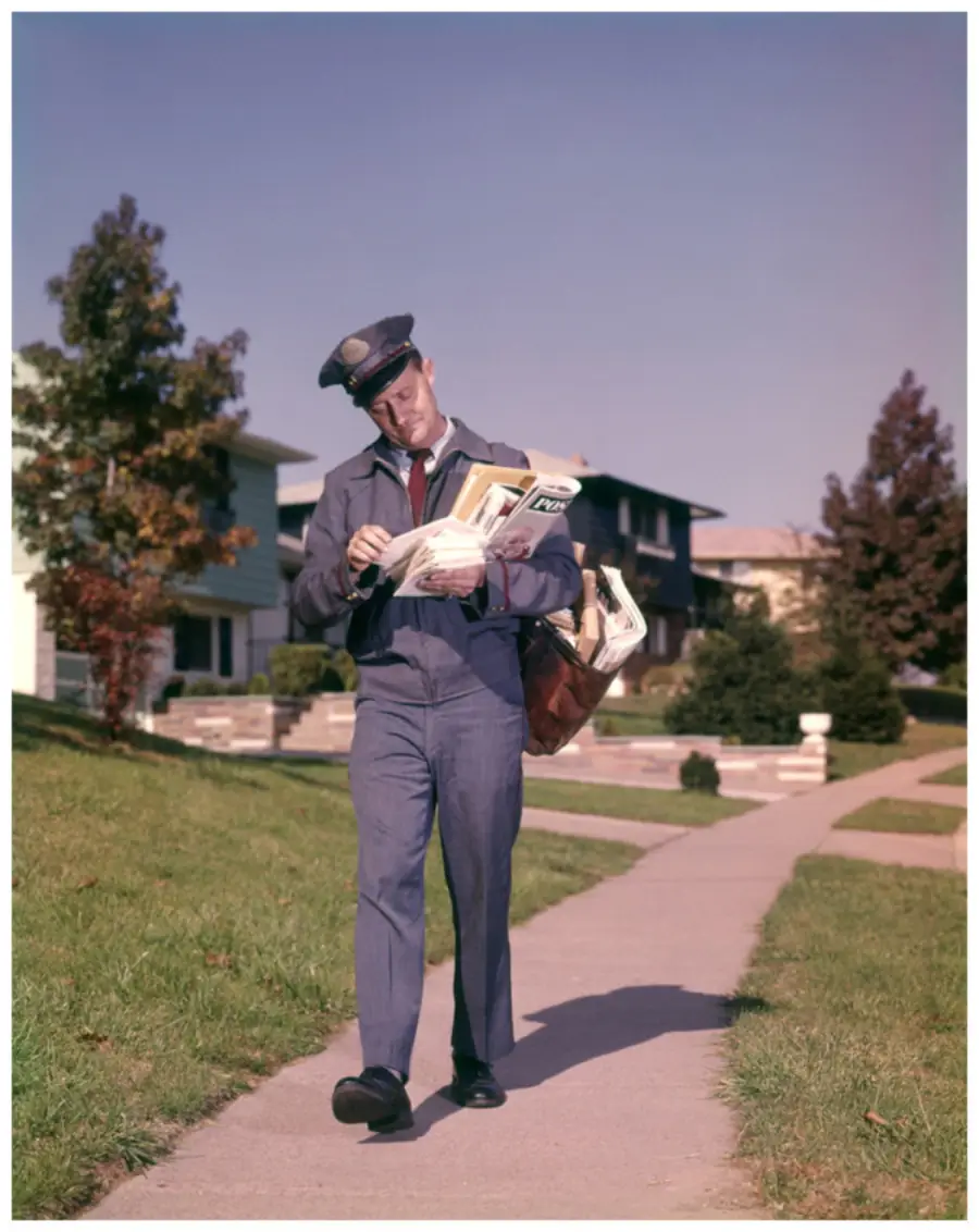 A mail carrier in a blue uniform walks on a suburban sidewalk, carrying a mailbag full of letters and packages while sorting mail. Houses and trees line the street on a sunny day.