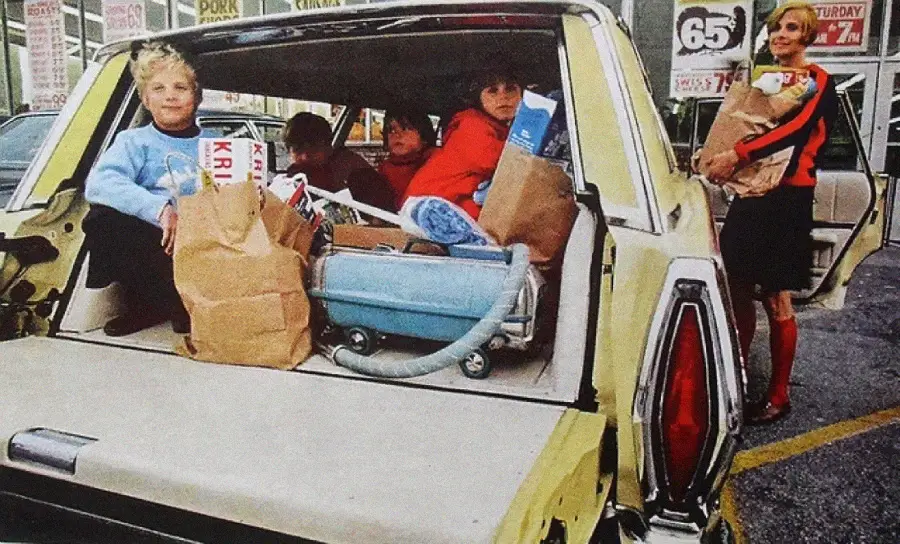 A woman stands by a car holding groceries while children sit in the back amid brown paper bags and a blue vacuum in a store parking lot.