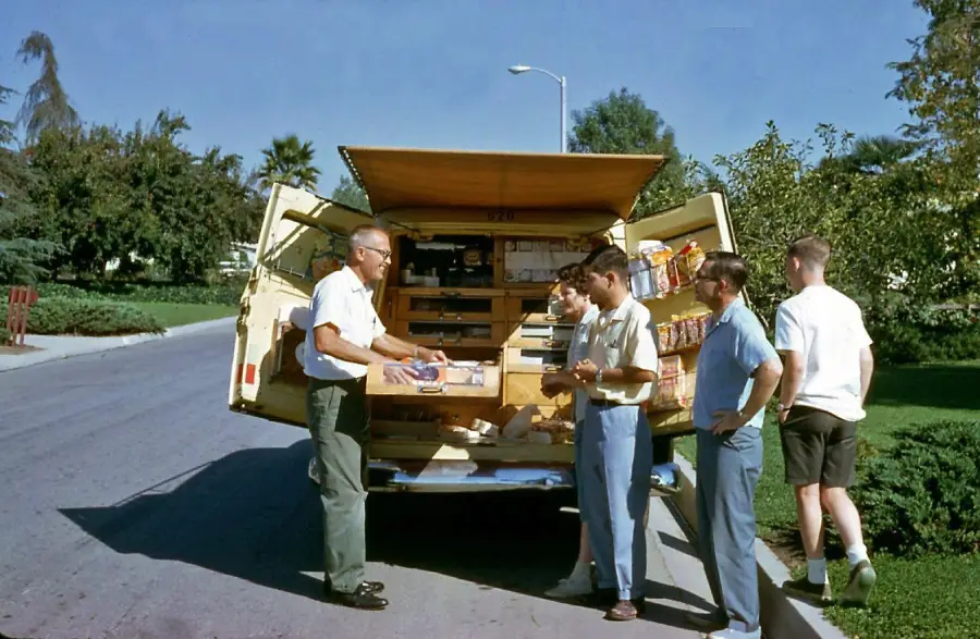 A man stands by an open van displaying snacks and goods to four young men on a sunny suburban street lined with trees and grass.