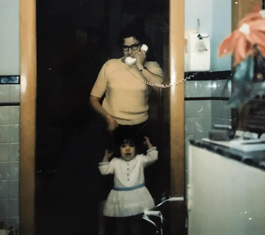 A woman stands in a doorway talking on a corded phone, while a young girl in a white dress with a blue belt stands in front of her, raising her hands by her head.