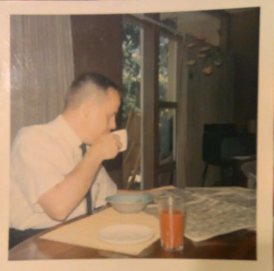 A man in a white shirt sits at a table, drinking from a cup. A bowl, a glass of juice, and a newspaper are on the table. Sunlight streams through windows in the background.