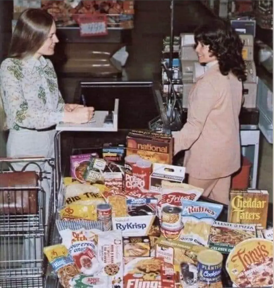 Two women stand at a grocery store checkout counter, talking. The shopping cart is filled with various colorful packaged foods, mostly snacks and breakfast items, from different brands.