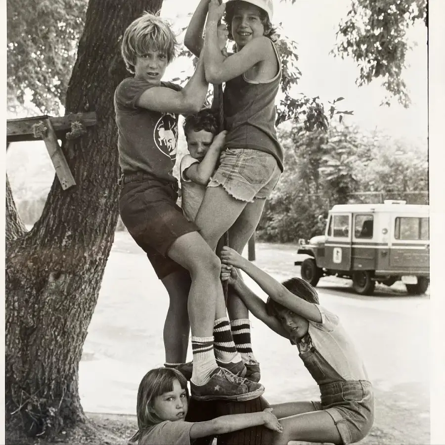 Five children play by climbing and hugging a wooden pole outdoors, with trees and a parked vehicle in the background. The photo is black and white.