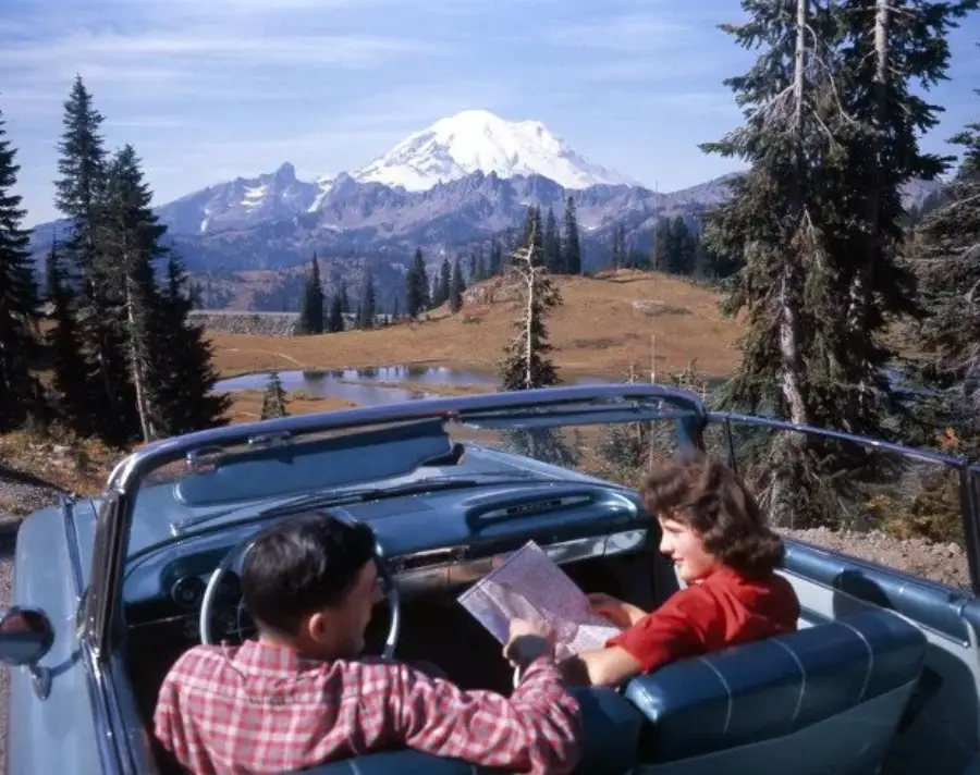 A couple sits in a blue convertible, looking at a map. Pine trees and a distant snow-capped mountain are visible beneath a blue sky.