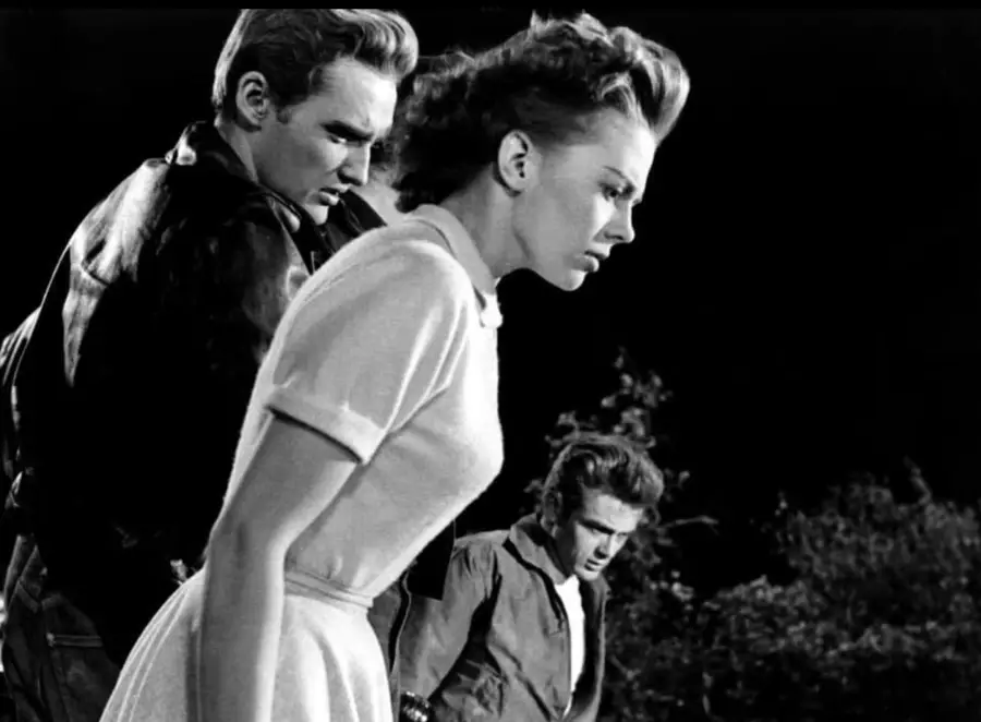 Three young adults stand outdoors at night, looking down with serious expressions. The woman in front wears a light dress, while the two men behind her wear jackets. Shadows and trees are visible in the background.