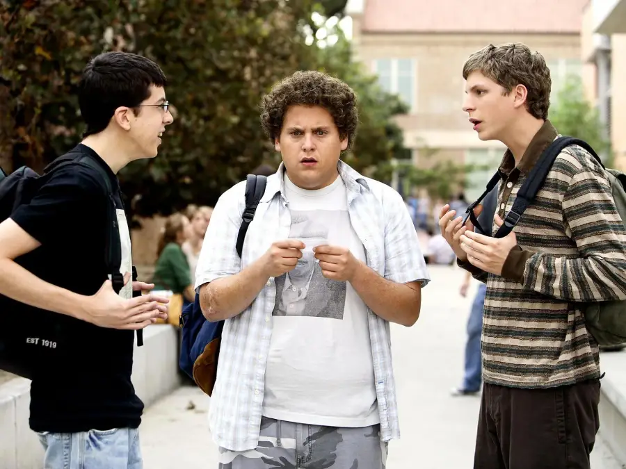 Three young men stand outdoors in a scene from the movie Superbad. Christopher Mintz-Plasse on the left, Jonah Hill in the center looking surprised, and Michael Cera on the right are having a conversation while wearing backpacks on a school campus.