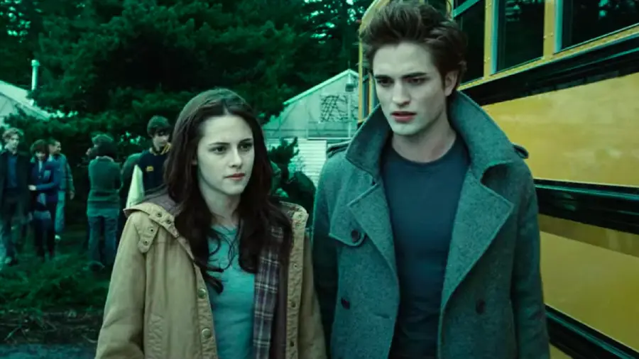 A young woman and a young man with pale skin stand beside a yellow school bus, looking serious. Other students are visible in the background near trees and a building.