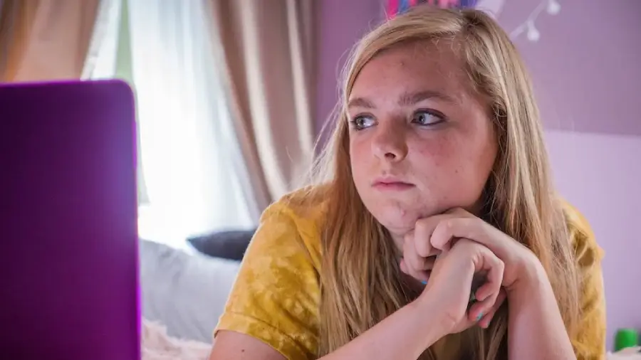 A young person with long blonde hair sits indoors, resting their chin on their hands, and looks thoughtfully toward a purple laptop screen. Light from a nearby window brightens the room.