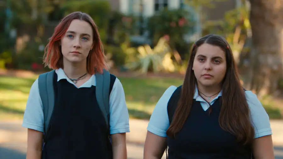 Two teenage girls in school uniforms stand outdoors, looking serious. One has shoulder-length red hair and a backpack, the other has long brown hair. Trees and a house are blurred in the background.