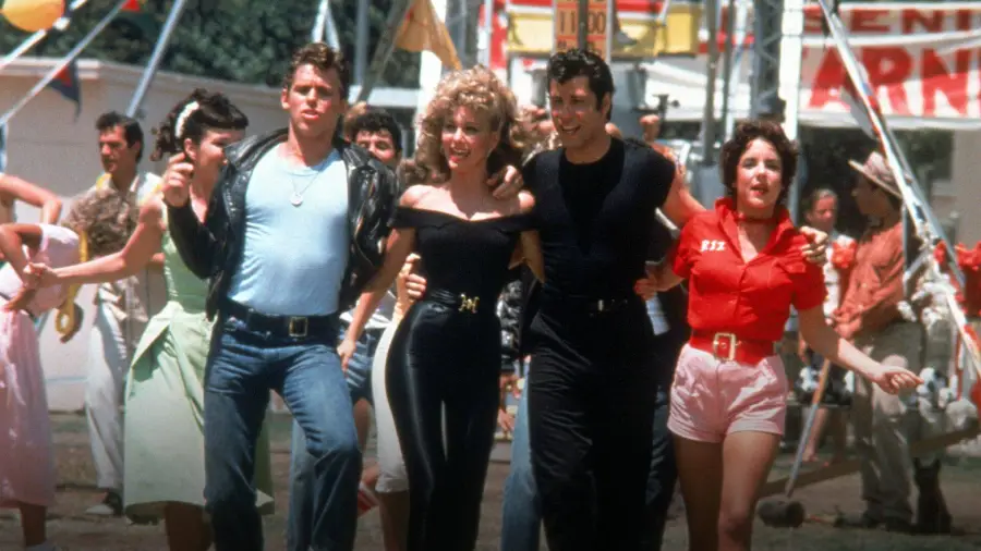 Four young adults walk arm-in-arm at a fair, dressed in 1950s-style outfits, smiling and singing. A crowd and carnival rides are visible in the colorful background.