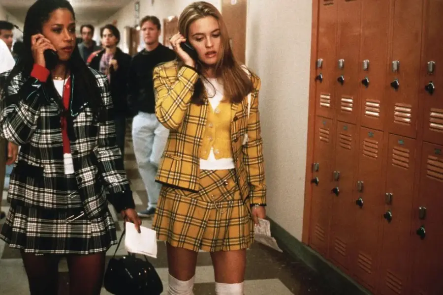 Two teenage girls in plaid skirts and blazers walk by lockers in a school hallway, talking on cell phones. One wears yellow plaid, the other black and white. Other students are visible in the background.