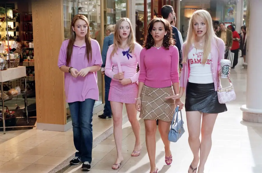 Four young women walk through a shopping mall. Three wear pink tops and skirts, while one wears a purple polo and jeans. They appear to be talking together. The setting is bright and busy, with stores in the background.