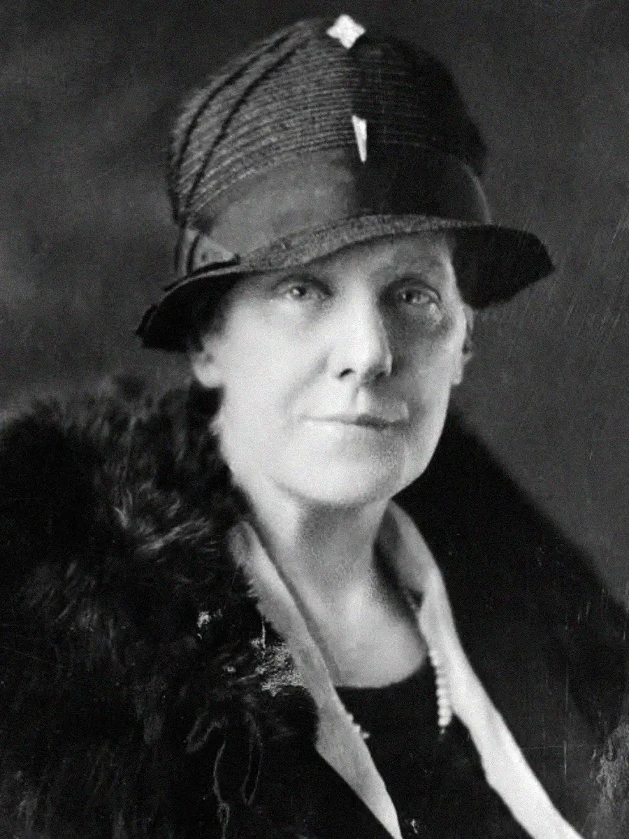 Black-and-white portrait of an older woman wearing a dark hat with a brooch, fur-trimmed coat, and a pearl necklace, looking directly at the camera with a neutral expression.