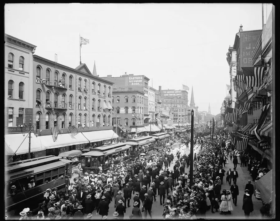 A busy city street scene from the early 1900s shows crowds of people, trolleys, and buildings with awnings and flags. The street is lively, filled with men, women, and children dressed in period clothing.