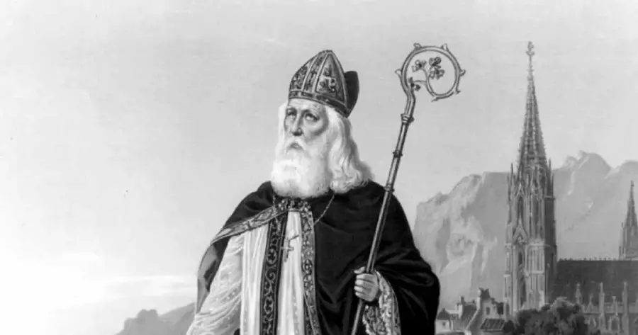 A bearded man in religious robes and a bishop’s hat holds an ornate staff, standing outdoors with a large gothic church and mountains in the background.